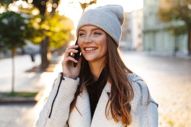 女性一个快乐美丽的红发女人戴着帽子走在户外用手机交谈的画面人女孩电话