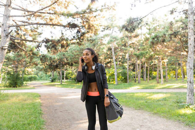 耳机20多岁的美女穿着黑色运动服 戴着耳机 在绿色公园用手机说话训练运动移动