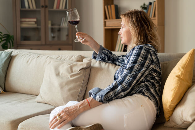 沙发严肃的金发熟女 穿着休闲装 在家里坐在沙发上 端着一杯红酒 看着饮料美女休息葡萄酒