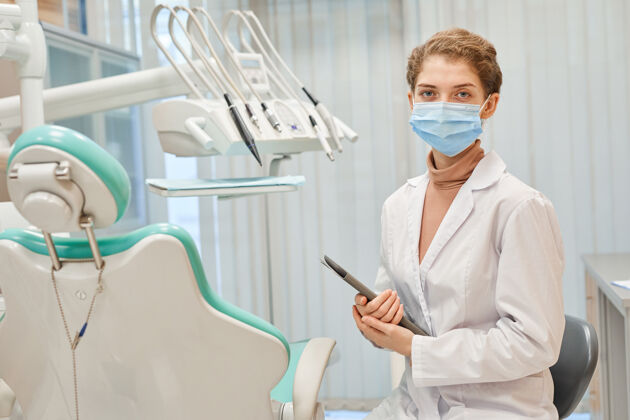 表情戴着口罩 穿着实验室工作服的年轻牙医的肖像 站在办公室里拿着数字平板看前面专业职业防护服室内