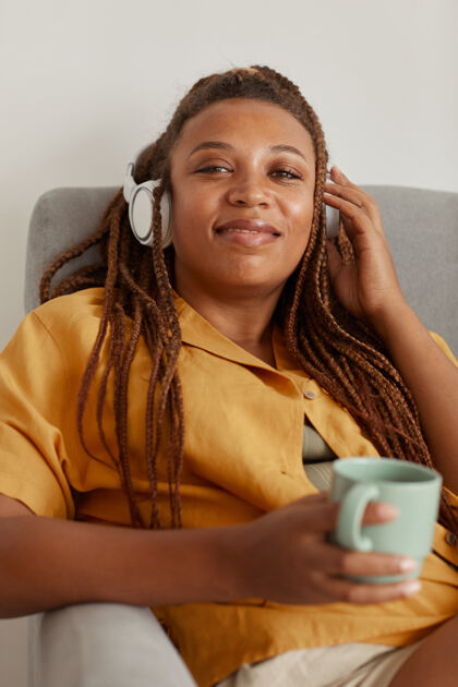 家庭内部戴着耳机的非洲妇女坐在扶手椅上喝着咖啡对着相机微笑的画像聆听非洲人后裔沙发
