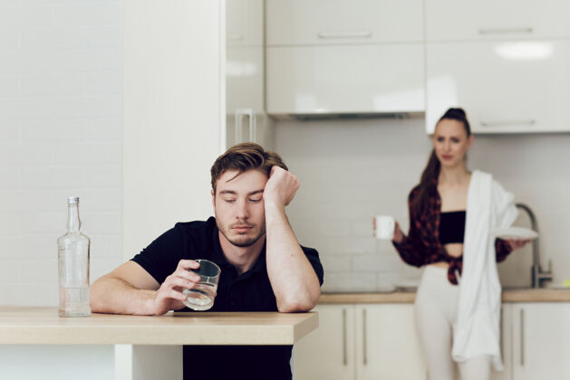 醉酒一个男人坐在厨房的桌子旁喝酒 一个女人在他后面吵架大叫家庭关系厨房