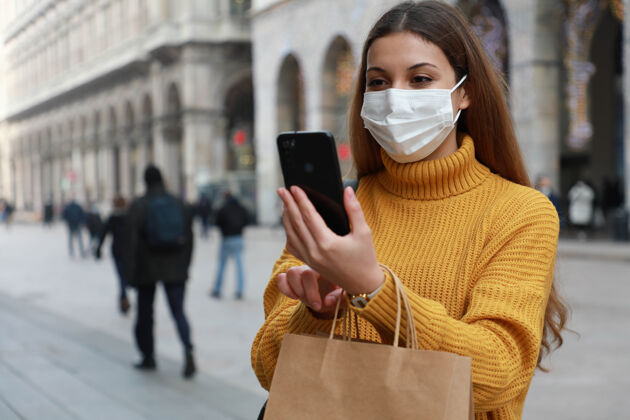 佩戴购物妇女与外科口罩使用智能手机应用程序在城市街道人女孩户外