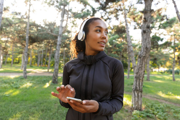 女性一位20多岁的女运动员穿着黑色运动服 戴着耳机 一边在绿色公园散步 一边用手机拍照成人听年轻人