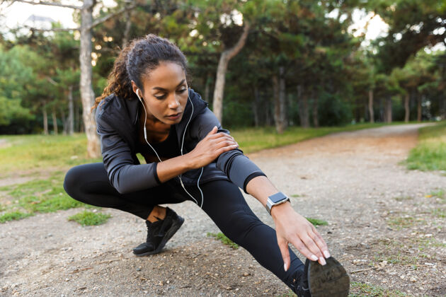 黑发一幅20多岁的非洲裔美国年轻女子穿着黑色运动服在绿色公园里锻炼 伸展双腿的画像户外吸引自然