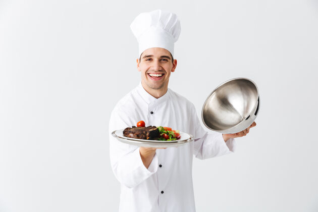 微笑兴奋的男厨师穿着厨师制服打开便当盖隔离在白色的墙壁上 展示肉菜欢呼烹饪烹饪