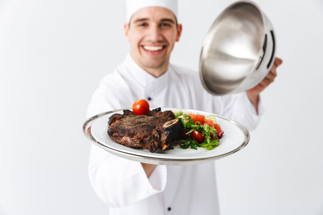 概念兴奋的男厨师穿着厨师制服打开便当盖隔离在白色的墙壁上 展示肉菜人厨具高加索