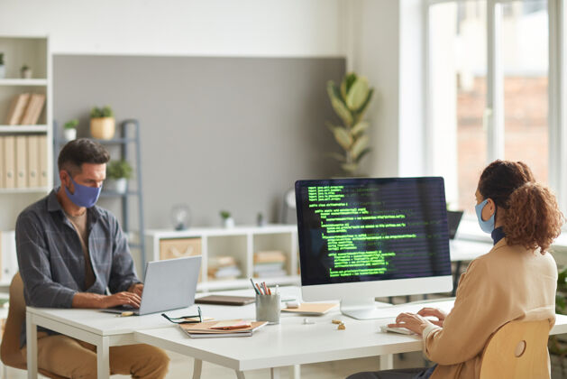 会议两个戴着防护面具的程序员在it办公室的桌子旁的电脑上编写软件和脚本商务女性互联网坐姿
