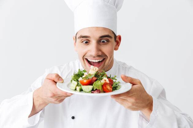 营养兴奋的男厨师穿着制服 把新鲜的蔬菜沙拉放在隔离在白色墙上的盘子上职业服务手