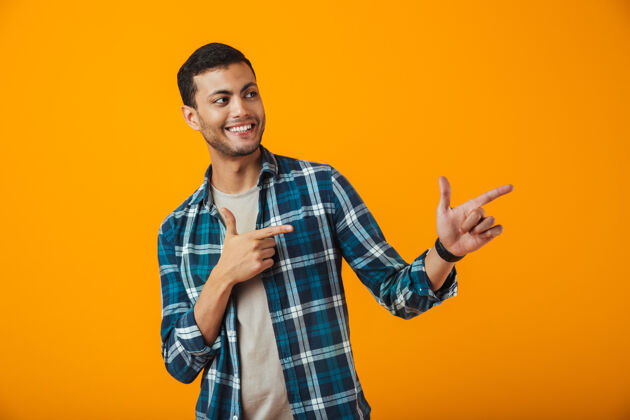 男孩一个穿着格子衬衫的快乐的年轻人孤零零地站在橙色的墙上 用手指着复制空间兴奋成人手势