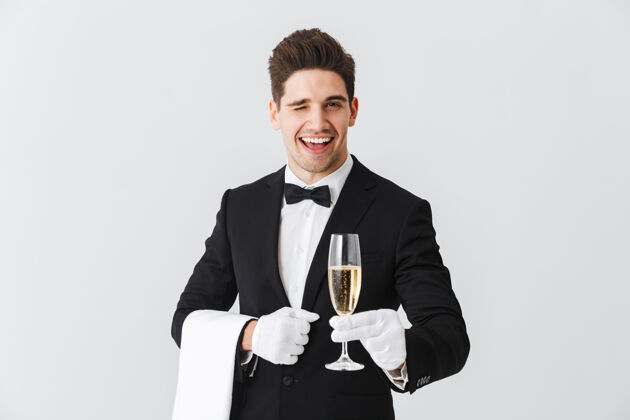 杯子身着燕尾服的微笑年轻侍者的画像在白墙上为您奉上一杯香槟手套液体香槟