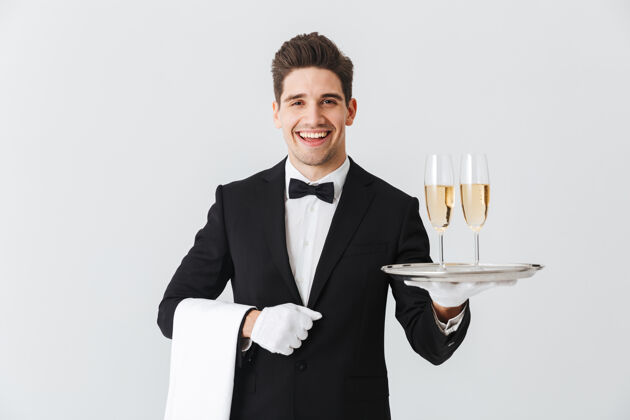 前面一个穿着燕尾服的微笑的年轻侍者的肖像 白色墙壁上放着两杯香槟男性工作饮料