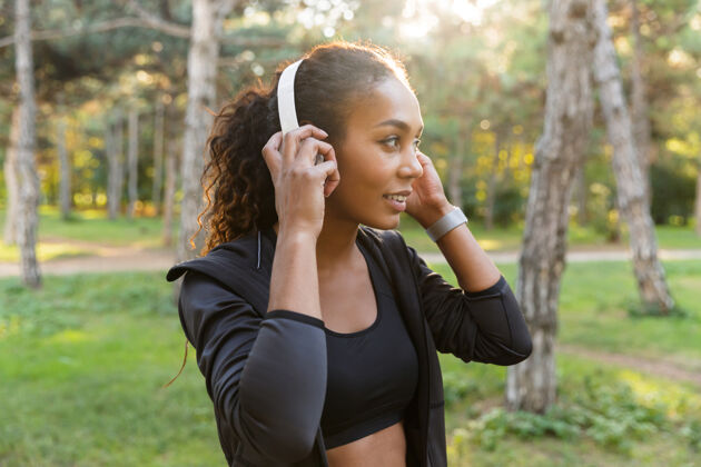 早晨20多岁的美女穿着黑色运动服 戴着耳机 穿过绿色公园美国女性女性