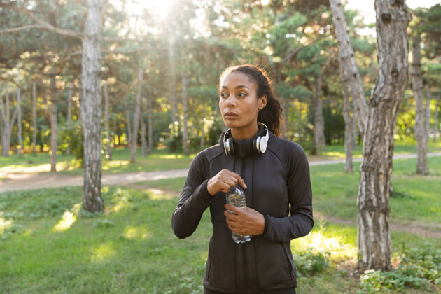 美国人20多岁的女运动员穿着黑色运动服 戴着耳机 拿着水瓶穿过绿色公园精力充沛身体深色头发