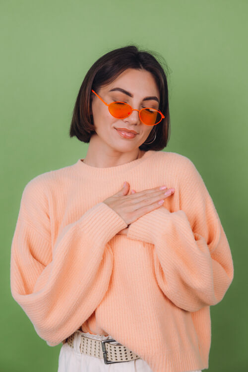 温柔年轻时尚的女士穿着休闲桃色毛衣 戴着橘色眼镜 隔着绿橄榄色的墙正面双手合十放在胸前 心复制空间情绪真诚室内