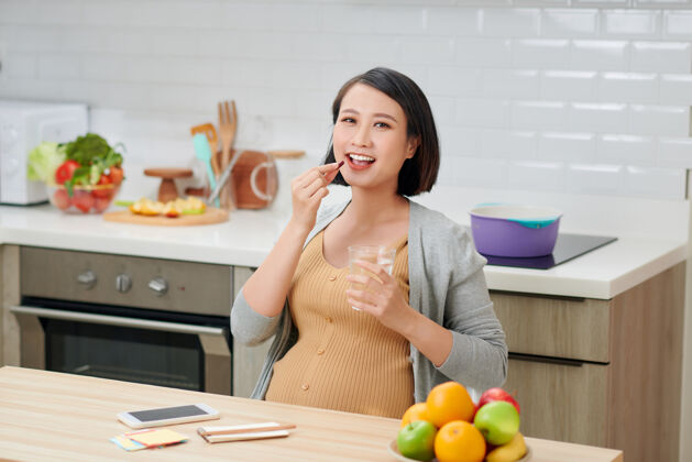 期望孕妇拿着维生素药片和一杯水放在厨房里孕妇女孩成人