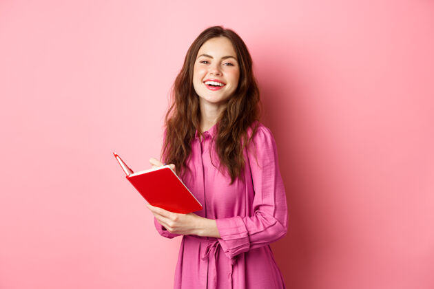 惊人美丽的年轻女子微笑着 写在笔记本上 拿着计划者或日记 站在粉红色的墙上水平微笑写作