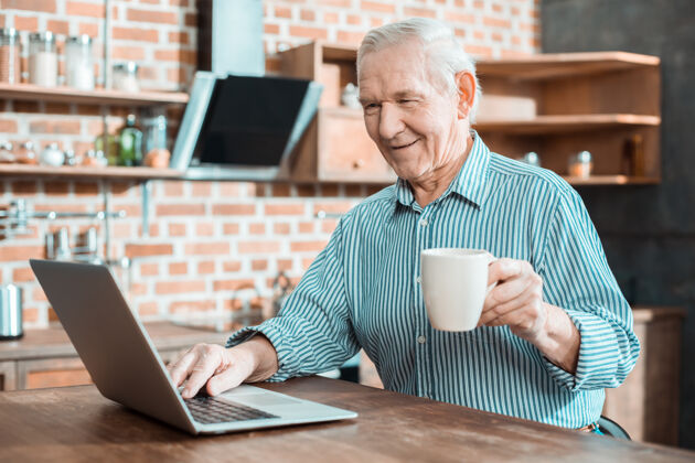 特写高兴的积极的老人微笑着拿着一杯茶 同时使用笔记本电脑退休放松茶