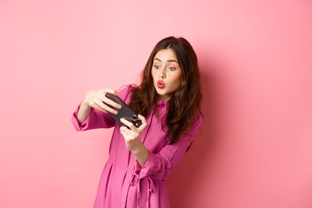 女性科技概念无忧无虑的年轻女子倾斜身体 水平地拿着智能手机 在手机上玩电子游戏 站在粉红色的墙上深色头发积极魅力