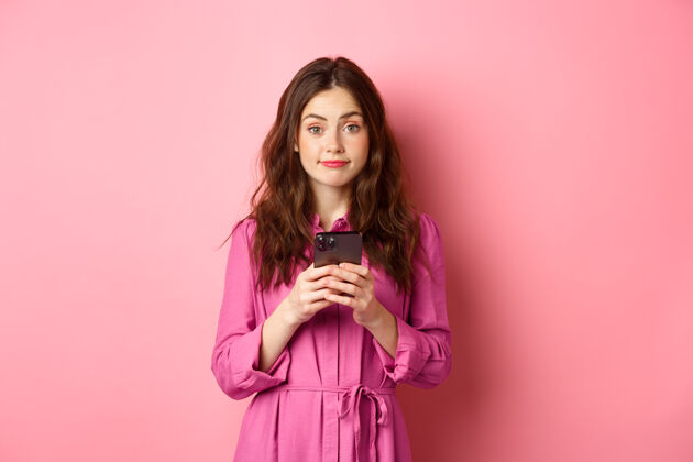 魅力可爱的年轻女子使用手机应用程序 手持智能手机 对着相机微笑 穿着裙子站在粉红色的墙上复制空间休闲赞异应用程序