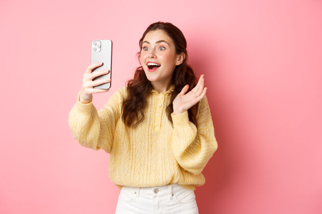 应用程序女人视频聊天惊讶的脸 收到惊人的消息 喘息兴奋 看到智能手机屏幕上的东西 站在粉红色的墙壁休闲在线魅力