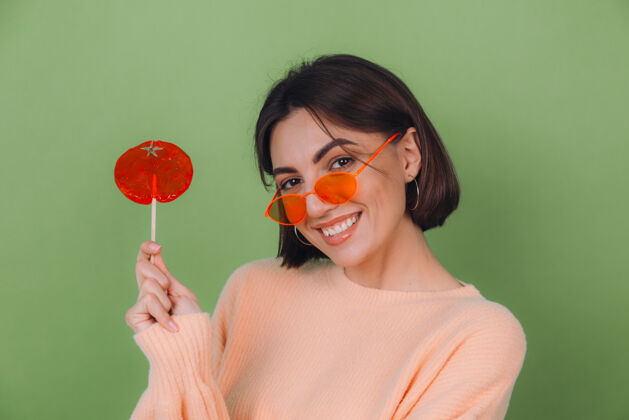 表情年轻时尚的女士穿着休闲的桃色毛衣 戴着橙色的眼镜 隔离在绿色橄榄色的墙上 带着橙色棒棒糖的正面微笑复制空间食物时尚无忧无虑