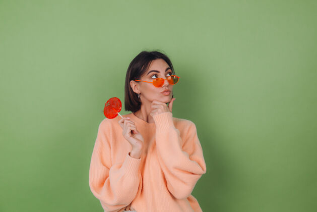焦糖年轻时尚的女士穿着休闲的桃色毛衣 戴着橘色眼镜 隔离在绿色橄榄色的墙上 用橘色棒棒糖若有所思地看着一边思考着复制空间无忧无虑持有可爱