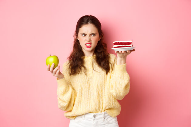 水平节食的健康女孩厌恶地看着一块蛋糕 拿着绿色的敷 做着不满意的鬼脸 站在粉红色的墙上积极乐观蛋糕