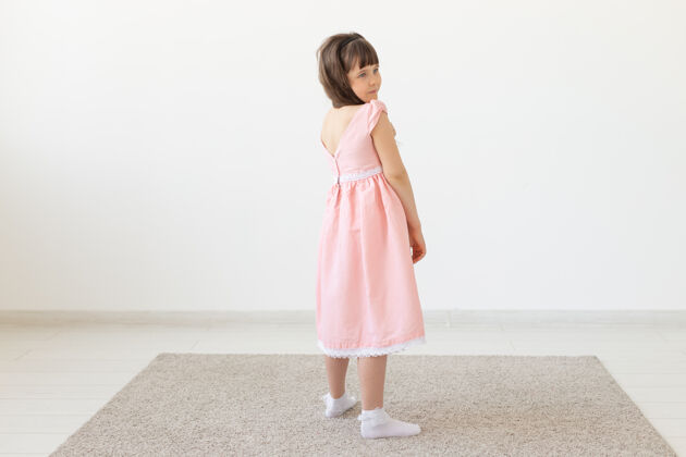 积极穿着粉色连衣裙的迷人小女孩微笑着 对着白墙的表面看着镜头可爱童年魅力