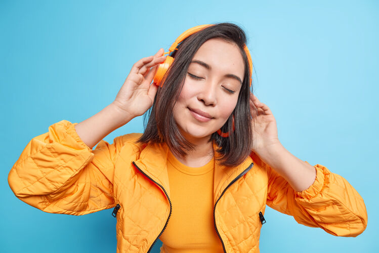 安静美丽的黑发女人闭着眼睛戴着无线耳机听音乐歪着头穿着橙色夹克隔着蓝色的墙人们的生活方式爱好概念高兴旋律学生