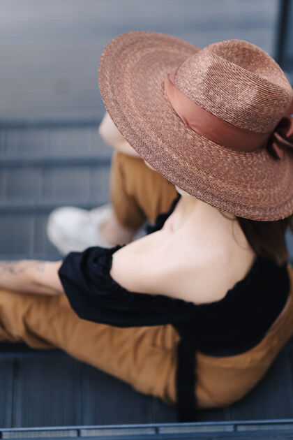 配件时髦的黑发女人留着长发 戴着时髦的大柳条帽 在楼梯上摆姿势城市时尚女人时尚