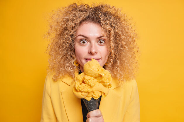 姿势惊讶的卷发女人吃着美味的冰激凌 不在乎卡路里 看着令人印象深刻的快乐表情 隔着黄色的墙壁女人的甜点是冰淇淋积极脸欧式