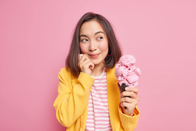 人梦幻的东方女人带着若有所思的表情看着远方 手里拿着美味的冰淇淋 想着什么 手放在脸上 穿着时髦的衣服 隔离在粉色的墙上举行夏天请