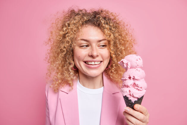 卡路里快乐的卷发女人的画像微笑着看着开胃的冰淇淋在黑色华夫饼吃美味的夏季甜点穿着时髦的服装感觉诱惑隔离在粉红色的墙上衣服女人人