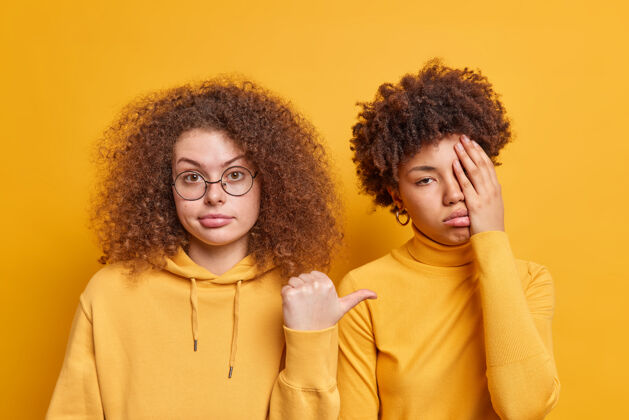 人美丽的卷发女人用大拇指指着她无聊沮丧的女性朋友 想知道为什么她穿着沮丧的衣服被隔离在黄色的墙壁上人们的情感和多样性的概念消极不满人