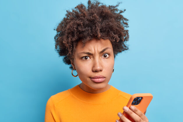 信息不满的非裔美国妇女微笑着脸看着不满的表情手持现代手机在线阅读负面新闻穿着橙色套头衫隔着蓝色的墙科技自负恼怒休闲傻笑