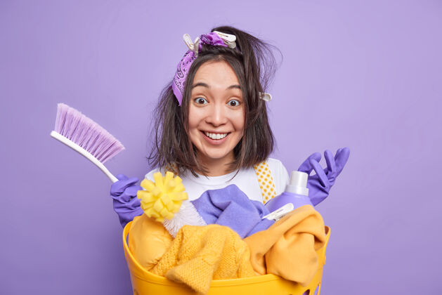 设备男人拿着衣夹在头发上 拿着刷子忙着打扫屋子 微笑着站在洗衣篮旁 用洗涤剂隔离在鲜艳的紫色上洗衣女除尘器