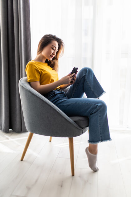 在线肖像年轻的亚洲妇女使用手机坐在椅子上客厅内部人科技杯子