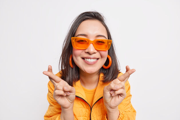乐观快乐时尚的深色头发的亚洲女人许下愿望祈求梦想成真微笑快乐地戴着时髦的橙色太阳镜耳环和夹克隔着白墙欢呼休闲成人