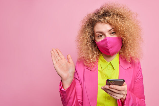 人困惑犹豫不决的卷发女人穿着鲜艳的正式衣服准备商务会议戴着防护面具在检疫检查新闻稿通过智能手机隔离在粉红色的墙上病毒穿着不知所措