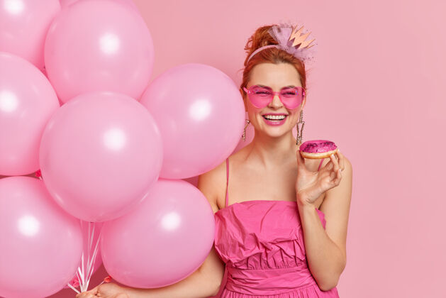 甜点积极时尚的红发女人笑容灿烂有喜庆的心情拿着美味的甜甜圈充气气球戴着心形太阳镜和连衣裙束生日阴影