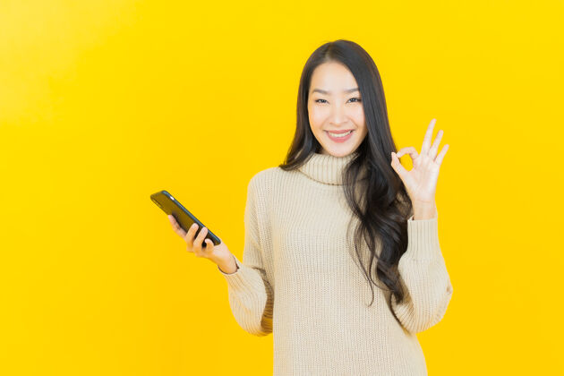 购物画像美丽的亚洲年轻女子微笑着在黄色的墙上智能手机美丽设备个人