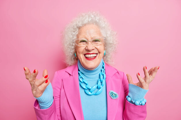 微笑快乐的高级卷发女士享受晚年生活举手微笑积极穿着时髦的服装化妆鲜艳有红色指甲皱纹的脸欢呼积极美丽