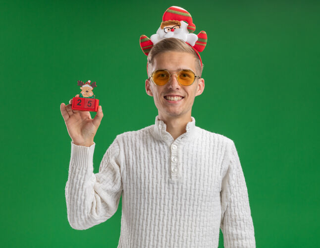 年轻人微笑着的年轻帅哥戴着圣诞老人的头带戴着眼镜拿着驯鹿玩具和日期看起来孤立在绿色的墙上微笑眼镜圣诞老人