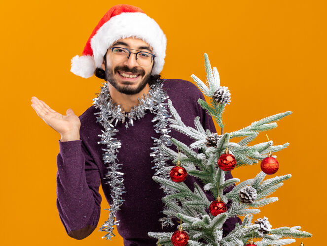 蔓延微笑的年轻帅哥站在圣诞树后面 戴着圣诞帽 脖子上戴着花环 手被隔离在橙色的墙上花环手帅气