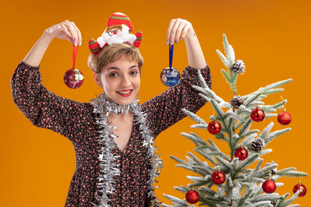 立场快乐的年轻漂亮女孩戴着圣诞老人的头带 脖子上戴着金属丝的花环 站在装饰好的圣诞树旁 拿着圣诞饰品 靠近孤立在橙色墙上的头圣诞老人花环女孩