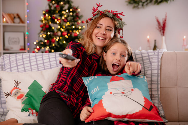 冬青带着冬青花环的微笑着的母亲拿着电视遥控器 坐在沙发上和女儿在家里享受圣诞节时光女儿坐着点