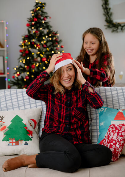 坐微笑的女儿把圣诞帽戴在妈妈头上 坐在沙发上 在家里享受圣诞时光沙发头享受