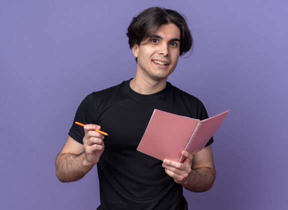 穿着面带微笑的年轻帅哥穿着黑色t恤拿着笔记本用钢笔孤立在紫色的墙上微笑男人感觉