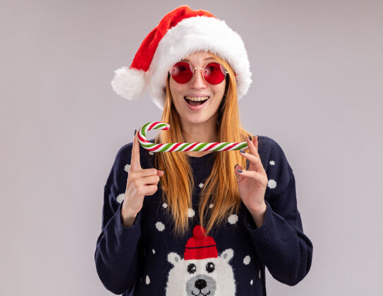 抱着微笑的年轻漂亮女孩戴着圣诞帽戴着圣诞眼镜拿着圣诞糖果孤立在白色的墙上眼镜年轻圣诞节